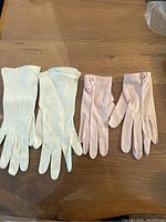 Front view of two pairs of vintage ladies gloves on wood surface, one cream and one light pink with button detail
