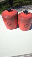 Photo showing two red vintage metal canisters with lids on white surface outdoors in sunlight. Both have black square knobs on their lids and vintage labels on front.