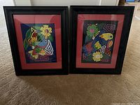Two abstract framed fabric art pictures positioned on floor showing hummingbird on left and parrot on right. Both have black frames with red matting and vibrant colored fabric designs.