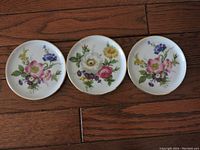 Top-down photo showing three small round ceramic plates arranged in a line on a wooden surface, each plate with a different floral design.