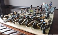 Display of multiple pewter battle figurines including soldiers, horses, and cannons arranged on wooden board.