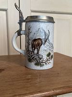 Front and side view showing ceramic stein body with painted mountain goat and scenic mountain background, crackle glaze visible, pewter lid with engraved stag, and metal thumb lift on handle.