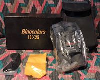 Photo showing RUKO 10x25 binoculars sealed in plastic bag with black carrying case, original black box with gold lettering "Binoculars 10x25", a yellow lens cleaning cloth, black lens caps, and a neck strap laid out on patterned fabric.
