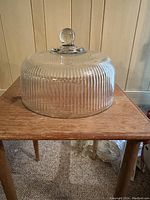 Ribbed clear glass cake lid with rounded handle knob placed on wooden table against wall background
