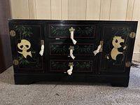 Front view of black lacquered dresser showing pandas on cabinet doors and drawers with metal flower-shaped knobs and white fabric tassels.
