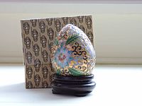 Photo showing the cloisonné enameled egg with blue flower and gold swirls on a dark wooden stand, alongside its original box with geometric pattern.