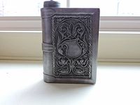 Front view of book shaped pewter flask standing upright on a white surface in natural light.