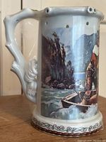 Porcelain dribble beer stein featuring a colorful scene of two men in a wooden boat on a river with rocky cliffs in the background. The stein is white with blue, green, and brown tones in the image. The handle is sculpted resembling a branch with leaf motifs. The top rim has multiple holes.