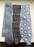 Three gray-tone men's silk ties on a light surface, showing geometric and intricate repeating patterns.