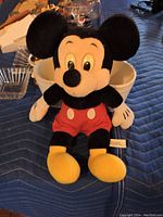 Mickey Mouse plush toy sitting upright on a blue quilted surface, showing front view with red shorts and yellow shoes.