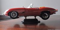 Side profile view of the red Jaguar E-Type model car displayed on a black stand.