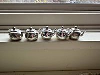 Five silver plated tea pot shaped name place card holders arranged in a row, showing the front and side details including handles, lids, and spouts.