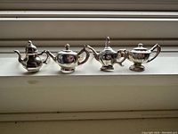 Front row of 4 tea pot shaped napkin rings displayed on a white surface under natural light, showing variations in design such as lid knobs, handle shapes, spouts, and ribbed textures.