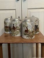 Four porcelain beer steins grouped on a wooden table with cast pewter lids, featuring four different wildlife designs including a deer, pheasant, rabbit and boar.