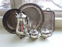 Photo showing the full silver set including three serving trays of various sizes and shapes, a tall tea pot, sugar bowl, and creamer, all arranged on a white surface in front of a window with snowy outdoor background.