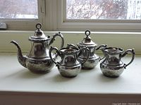 Full view of the 4-piece silver tea set including teapot, sugar bowl, and two creamers placed on a white surface in front of a window.