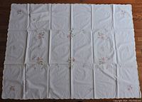 Full view of rectangular white tablecloth with scattered pink, purple, and yellow floral embroidery on fabric laid flat to show entire design and scalloped edges.