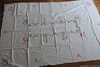 White rectangular tablecloth adorned with pink flowers and cutwork lace, laid out flat on wooden floor.