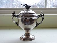 Single photo showing the entire silver sugar bowl with raised pedestal base, two ornate Victorian-style handles, and a lid topped with a bird figure. The bowl has a rounded, bulbous shape with polished silver surface reflecting its surroundings.