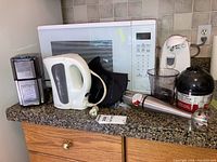 Full view of kitchen counter showing Cuisinart coffee grinder, electric kettle, hand blender with blending container, electric can opener, and black plastic food processor bowl. Microwave in background not included.