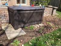 Full view of black plastic garden storage box placed outdoors next to a brick wall and garden area.