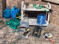 Photo showing white plastic two-shelf garden cart, blue watering cans, green watering bucket, garden hose attachments, lawn sprinklers, and spool of blue nylon twine.