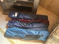 Four folded camp chairs in carry bags of different colors stacked on floor next to shelving unit.