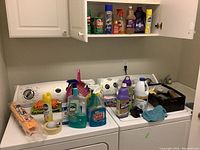 Cleaning supplies and containers arranged on top of washing machines and inside open wall cabinet.