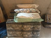 Photo of 8 outdoor chair cushions arranged on a surface with 4 bed pillows stacked on top, showing variety of colors and patterns