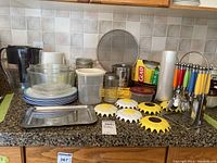 Photo showing various kitchen items including plastic plates, plastic bowls, water pitcher, glass sugar jar, and other containers and jars on a counter surface.