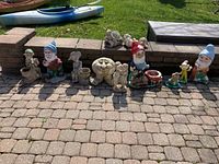 Wide shot showing all garden figures lined up on patio paving stones including gnomes, child figures, frog, and deer mostly in concrete and some plastic.
