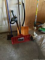 Shot showing multiple garden tools including snow shovels, hedge trimmer, grass trimmer and shovels grouped by a door.