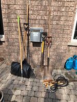 Full view of garden tools including two shovels, a snow shovel, a garden rake, hand saw, extension cord, and watering cans on side