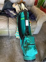 Photo of green golf bag containing multiple golf clubs including vintage wood drivers and irons, placed on a sagging sofa in a garage with various items in background.