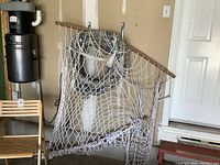 Full view of white netted full size hammock with wood spreader bars on each end hanging in garage near chair and wall hooks.