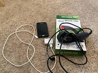 Adesso NuScan 2100 barcode scanner with box, Samsung smartphone, and white charging cable with plug visible on carpeted floor.