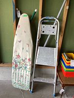 Ironing board with floral and butterfly patterned cover leaning against wall next to aluminum two-step stool.