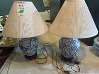 Two ceramic lamps with beige pleated shades visible from front showing cords, wooden bases, and floral design.