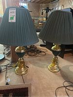 Pair of table lamps shown with pleated dark fabric shades, ornate brass-tone and green ceramic bases, placed on floor with cords visible.
