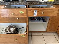 Open drawers and cupboard showing metal frying pans, baking pans, and metal colander stacked inside lower cabinet shelves.