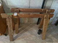 Photo showing two wooden saw horses standing upright in a basement with concrete floor and wall, showing wear and paint marks.