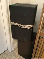 Two black rectangular tower speakers stacked with wires wrapped around them, showing the full shape and approximate size.