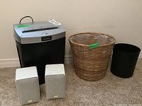 Pair of white Optimus Pro X-44AV speakers, Fellowes P-71 shredder, woven basket garbage can, and black plastic garbage can on carpeted floor against wall.