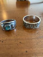 Photo showing two silver rings on wooden surface, left ring has turquoise stone, right ring has embossed design.