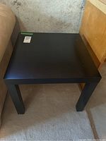 Black IKEA Lack side table positioned on carpeted floor beside furniture, showing tabletop and legs with visible scratches and nicks on surface.