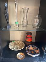 Collection showing wooden figurine, three clear glass vases, pottery vase, ceramic and metal bowls on two shelves.