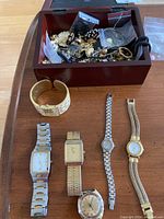 Overview of watches laid out on a wooden surface with a wooden box containing assorted jewelry pieces and plastic bags.