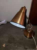 Photo of a brass table lamp with a conical shade, flexible neck, and round base on a floor surface.