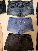 Front view of three pairs of women's American Eagle jean shorts showing different washes and details: medium blue distressed, light blue raw hem, and black faded pair.