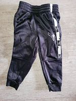 Photo showing a single pair of black baby pants with elastic waistband, cuffed ankles, and white Puma logos on side stripe, laid flat on a surface.